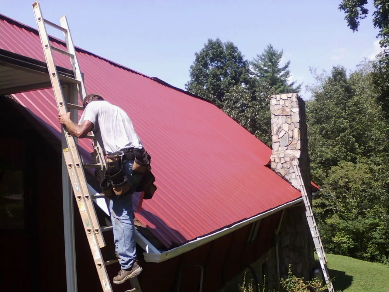 Roofer on a ladder beside a red metal roof and stone chimney — Soffit & Fascia Repair by Roofing Brothers in Southborough